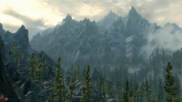 A screenshot from Elder Scrolls V: Skyrim. It's of mountains covered in pine forests, with wispy clouds in the sky.